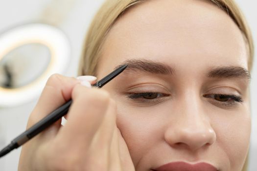 Stylist draws eyebrows with a pencil for a young beautiful woman.