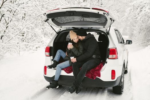 Young couple, a man and a woman, are sitting in the trunk of a car in a winter, snowy forest, hugging, kissing and drinking coffee.