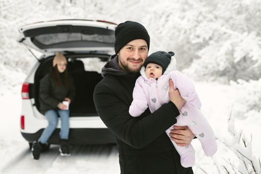 Young father holds a baby daughter in his arms against the background of a car in a winter snow-covered forest.