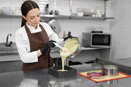 Young female pastry chef pours liquid mousse into a mold. Preparation of cake, dessert in a professional kitchen.