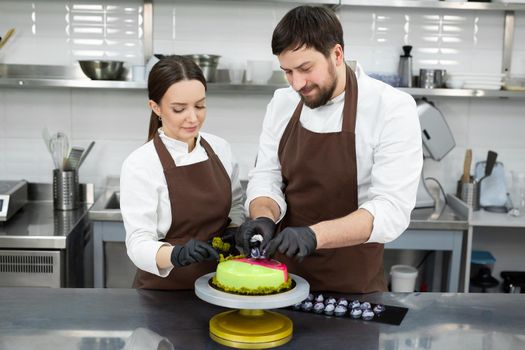 Two confectioners, a man and a woman, decorate a mousse cake with a mirror glaze with handmade chocolates.