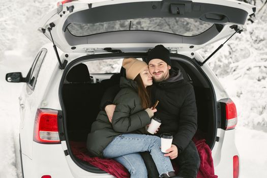 Young couple, a man and a woman, are sitting in the trunk of a car in a winter, snowy forest, hugging, kissing and drinking coffee.
