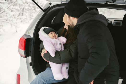 Young, happy family: a man, a woman and a baby are sitting in the trunk of a car in a winter snow-covered forest.