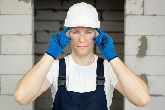 Handyman in a white T-shirt, a combination jacket and a hard hat puts on safety glasses.