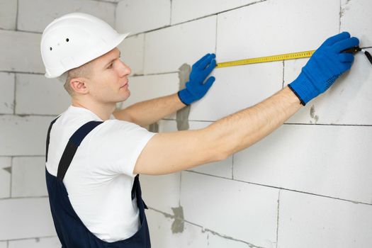 Modern engineer, a builder in overalls and a helmet measures the length of the wall of unfinished construction