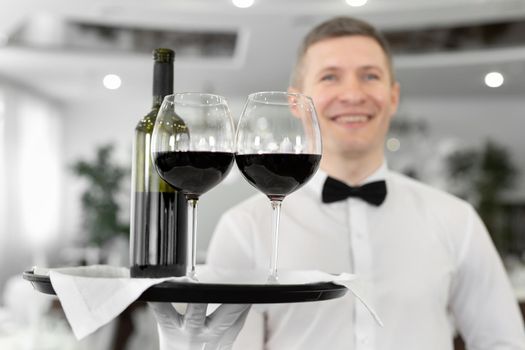 Smiling male waiter with glasses of red wine and a bottle on a tray in a restaurant.