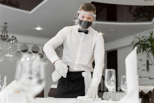 Portrait of a male waiter wearing a face mask and face shield, cleaning a table in a cafe during social distancing