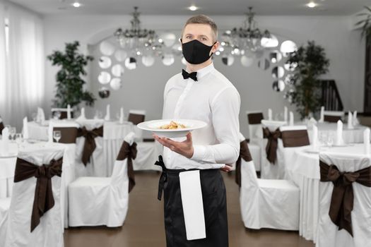 Young happy waiter wearing protective face mask while serving food in a restaurant.