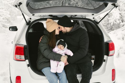 Young, happy family: a man, a woman and a baby are sitting in the trunk of a car in a winter snow-covered forest.