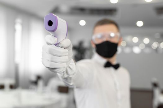 Waitress man wearing a face mask and holding an infrared thermometer on her forehead to check the body temperature for symptoms of the virus