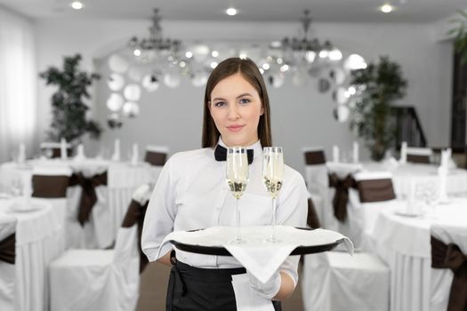 Happy female waiter with two glasses of white wine on a tray in a restaurant.