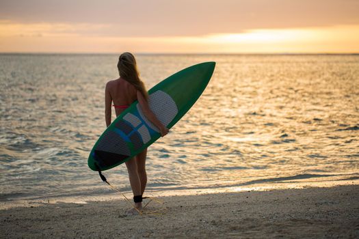 Surfing surfer girl looking at ocean beach sunset. Silhouette of female bikini woman looking at water with standing with surfboard having fun living healthy active lifestyle. Water sports with model.