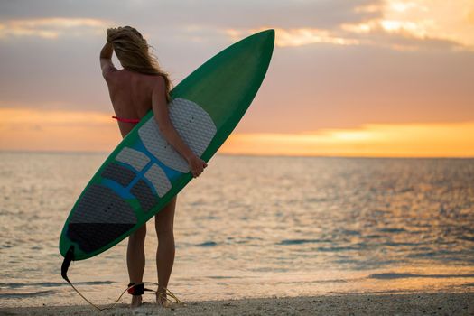 Surfing surfer girl looking at ocean beach sunset. Silhouette of female bikini woman looking at water with standing with surfboard having fun living healthy active lifestyle. Water sports with model.