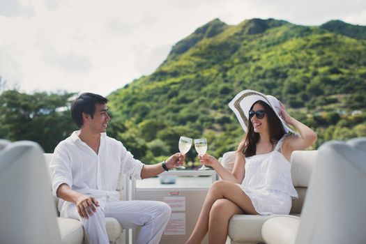 Smiling young couple holding glasses with champagne and looking at each other while sitting on the board of yacht