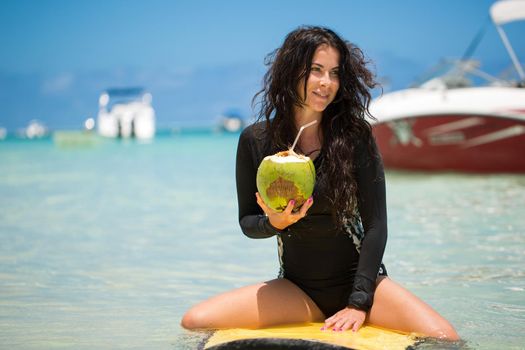 Portrait of a beautiful surfing girl with green coconut from palm tree sit on yellow surf longboard surfboard board