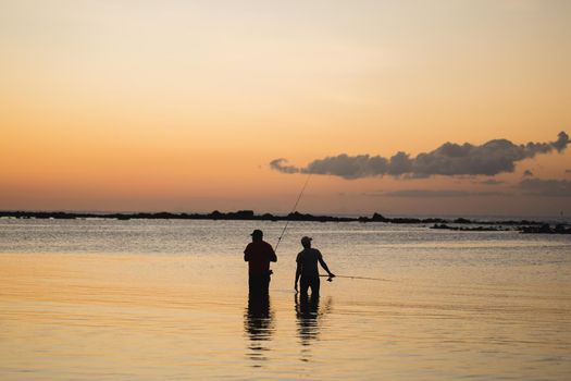 Two men fishing in the ocean from the beach at sunset