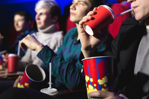 Glasses with a tube and popcorn in the hands of people in a movie theater close-up.