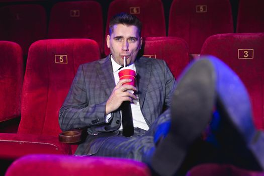 Portrait of a young, handsome man alone in a movie theater in a business suit, with his feet on the front seat and drinking through a tube from a red Cup.