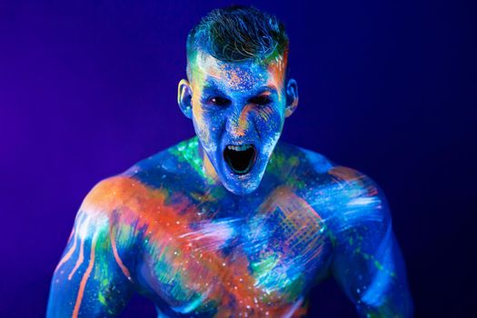 Young man shouts. Fluorescent paint on face and muscular torso, in studio shot with UV light