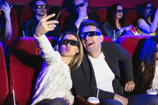 Beautiful girl and guy laughing happily making a selfie together during a movie at the cinema. Nice and positive girl and guy are taking selfie