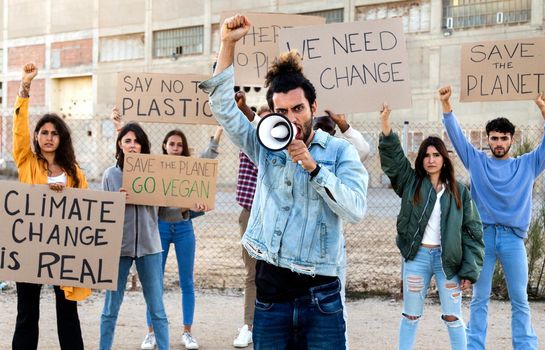 Angry young Arab man with arm raised shouting through megaphone protesting against climate change with group of demonstrators holding signboards. Activism concept.