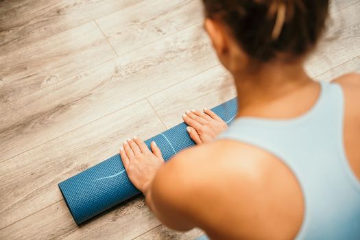 Woman hands rolled up yoga mat on gym floor in yoga fitness training room. Home workout woman close up hands rolling foam yoga gym mat. Woman barefoot home workout sportive healthy lifestyle concept.