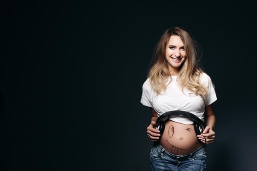 Happy and smiling pregnant female wearing white t shirt and jeans with big earphones on tummy. Expectant mother painting funny smile on her naked belly. Concept of maternety and health care.