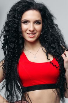 Front view of attractive long curly haired girl in sports red top smiling at camera. Sexy and hot dancing woman with perfect body sedactive posing. Concept of sport, dance, helthy lifestyle.