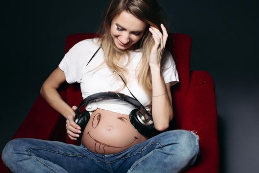 Happy and smiling pregnant female wearing white t shirt and jeans with big earphones on tummy. Expectant mother painting funny smile on her naked belly. Concept of maternety and health care.