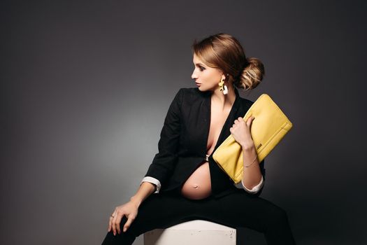 Portrait of elegant pregnant woman wearing in black suit with pregnant bell looking under jacket. Brunette girl holding yellow handbag, sitting against black studio background and looking at side.
