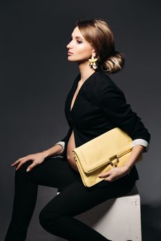 Portrait of elegant pregnant woman wearing in black suit with pregnant bell looking under jacket. Brunette girl holding yellow handbag, sitting against black studio background and looking at side.