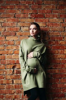 Portrait of happy pregnant brunette girl posing against brick wall. Stylish woman wearing in khaki dress, holding pregnant belly by hands, looking at side. Concept of pregnancy and paternity.