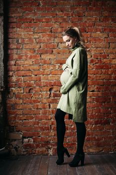 Portrait of happy pregnant brunette girl posing against brick wall. Stylish woman wearing in khaki dress, holding pregnant belly by hands, looking at side. Concept of pregnancy and paternity.