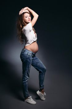 Beautiful pregnant lady wearing jeans and white t- short posing in studio. Future mother blowing kiss, looking at camera and showing her naked tummy. Concept of maternity. Black background.