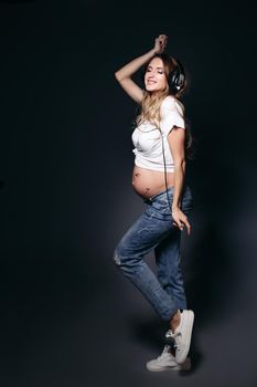 Beautiful pregnant lady smiling and posing with closed eyes. Expectant mother holding hand near head, listening music and showing naked tummy. Lady wearing in white tee shirt and blue jeans.