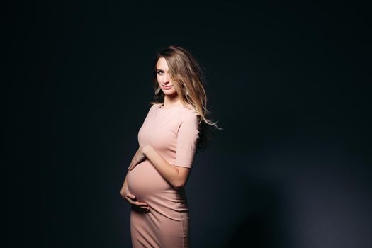 Windy effect on studio portrait of beautiful pregnant woman in pink dress, embracing belly. Elegant and stylish female posing, looking at side. Concept of pragnancy fashion and look.