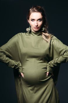 Front view of beautiful and fashionable pregnant brunette woman at dark studio. Pretty and stylish woman wearing in khaki dress, holding pregnant belly, looking at camera. Concept of pregnancy.