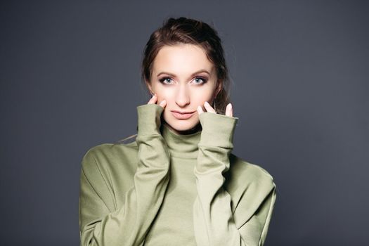 Close up of young brunette woman with big blue eyes, plump lips, wearing in khaki dress posing at studio, touching skin. Woman with perfect make up touching face by fingers, looking at camera.