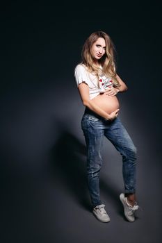 Front view of pretty and fashionable pregnant woman wearing in jeans and white t shirt embracing stomach. Future mother smiling at camera,holding hands on her naked tummy. Concept of pregnant fashion.