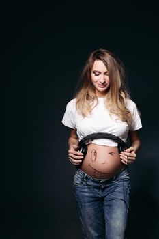 Happy and smiling pregnant female wearing white t shirt and jeans with big earphones on tummy. Expectant mother painting funny smile on her naked belly. Concept of maternety and health care.