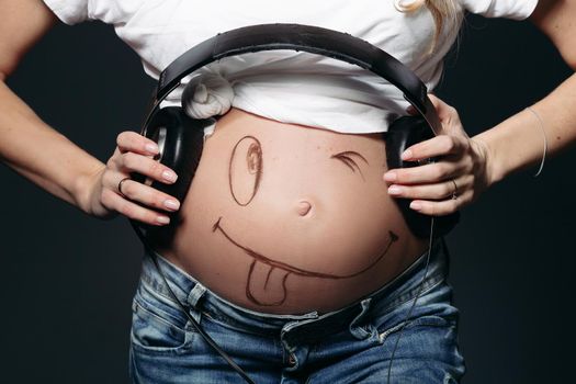 Close up of pregnant lady wearing white t-shirt, holding black earphones on tummy. Expectant mother painting funny smile on her naked belly. Concept of maternety and health care. Black background.
