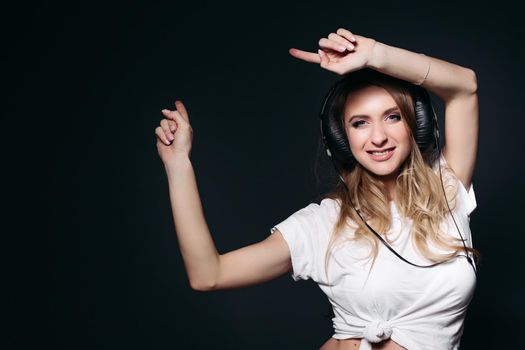 Happy and young pregnant mom dancing, smiling at camera and rising up by hands. Beautiful expectant mother listening music by big earphones and holding arms up. Female expacting her child.