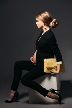 Portrait of elegant pregnant woman wearing in black suit with pregnant bell looking under jacket. Brunette girl holding yellow handbag, sitting against black studio background and looking at side.