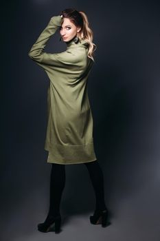 Front view of beautiful and fashionable pregnant brunette woman at dark studio. Pretty and stylish woman wearing in khaki dress, holding pregnant belly, looking at camera. Concept of pregnancy.