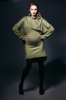 Front view of beautiful and fashionable pregnant brunette woman at dark studio. Pretty and stylish woman wearing in khaki dress, holding pregnant belly, looking at camera. Concept of pregnancy.