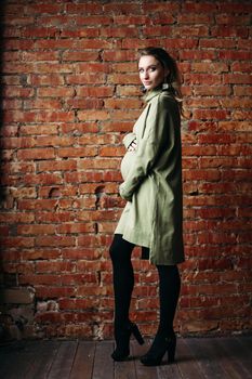 Portrait of happy pregnant brunette girl posing against brick wall. Stylish woman wearing in khaki dress, holding pregnant belly by hands, looking at side. Concept of pregnancy and paternity.