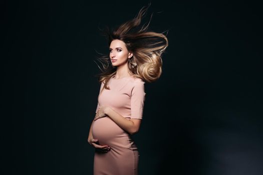 Windy effect on studio portrait of beautiful pregnant woman in pink dress, embracing belly. Elegant and stylish female posing, looking at side. Concept of pragnancy fashion and look.