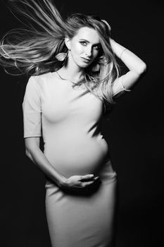 Black and white portrait of beautiful mom embracing stomach, smiling, looking away, over shoulder. Young happy mother posing at studio, against dark background. Pregnant fashion.