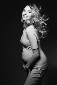 Monochrome portrait of beautiful, happy pregnant woman in dress, embracing belly and smiling. Positivity and stylish female posing, looking at camera. Concept of pragnancy fashion. Windy effect.