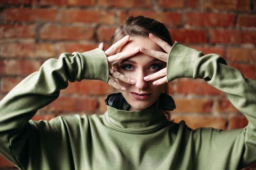 Close up of young brunette girl with big blue eyes, plump lips, wearing in khaki dress posing against brick wall. Woman with perfect make up touching face by fingers, looking at camera.